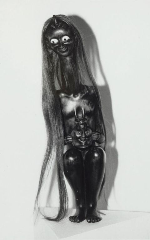 Sculpture. Black and white photograph of a sitting woman with a serpentine neck. Her head angles forwards and to our left, long hair affixed to the crown falling to her feet. Her bulging eyes are white with black pupils. The rest of her body is polished black.
