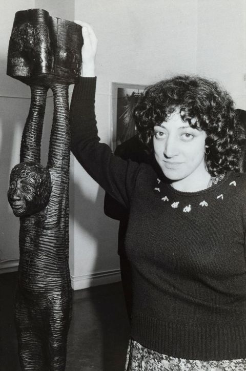Black and white photograph. A woman with black curly hair falling her shoulders stands next a sculpture as she looks at us. The sculpture is of a person with their arms raised. She rests one hand on the top, which comes up over her head.