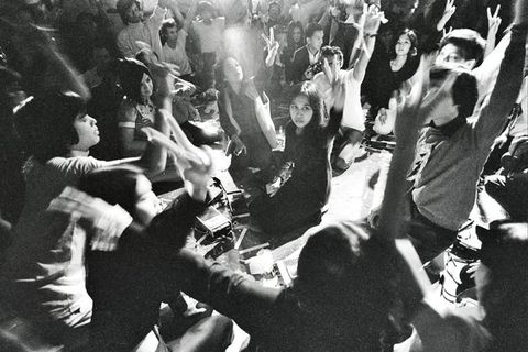Black and white photograph. About thirty-five people sit or kneel and hold up one hand, first and middle fingers raised to make a V. Cassette players sit on the ground between them.